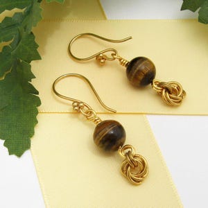 May include: Gold earrings with a brown tiger's eye gemstone and a gold knot design.