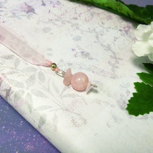 May include: A pink gemstone bookmark with a ribbon. The bookmark features a round rose quartz bead and two smaller, irregular-shaped rose quartz pieces, all attached to a silver wire. A pink ribbon and a gold bead complete the design.