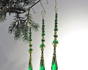 May include: Three Christmas ornaments with emerald green crystal pendants and gold-colored filigree and accents. The ornaments are suspended from gold-colored hooks and are decorated with green beads. The ornaments are hanging from a green pine branch.