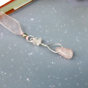 May include: A pink gemstone bookmark with a silver wire design. The bookmark features a teardrop-shaped rose quartz pendant and small rose quartz chips. A sheer pink ribbon is attached to the top, along with a silver bead.