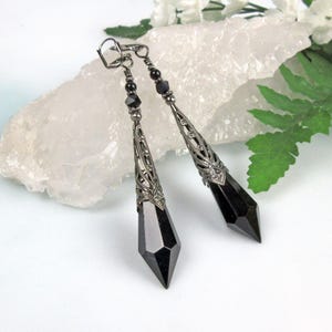 May include: A pair of long, black crystal drop earrings with silver filigree accents. The earrings are suspended from silver hooks.
