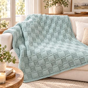 May include: A light mint green knitted throw blanket featuring a textured basketweave pattern with alternating vertical and horizontal stitch blocks, draped over the arm and seat of a neutral-colored upholstered sofa.