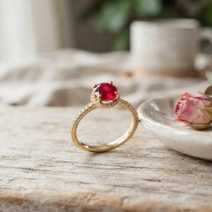 以下が含まれることがあります： 中央に鮮やかな赤色の丸い宝石がセットされ、その周りを小さな透明な石の輪が囲んでいるゴールドの指輪。バンドは細い金属製で、中央の赤い石を引き立てる光沢のあるゴールド仕上げが施されています。