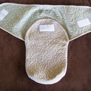 May include: A beige quilted baby wrap with a green quilted outer layer. The wrap has two white velcro closures on the outer layer and a white velcro closure on the inner layer.