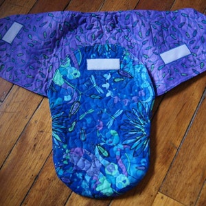 May include: A quilted baby carrier with a blue and purple floral print. The carrier has two white velcro straps on each side.
