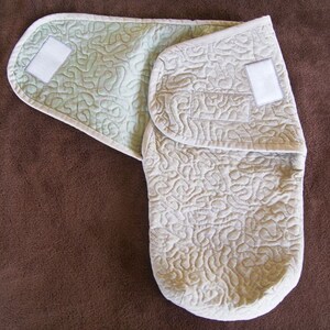 May include: A beige quilted baby swaddle with a green lining and two white velcro closures. The swaddle is folded in half and lying on a brown surface.