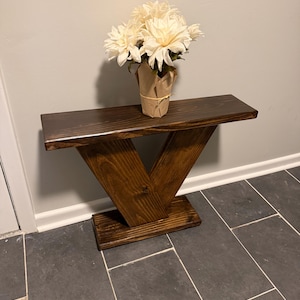 May include: A dark brown stained wooden console table with a V-shaped base and a rectangular top. A bouquet of white flowers wrapped in brown paper sits on the surface of the table. The wood grain is visible throughout the structure.