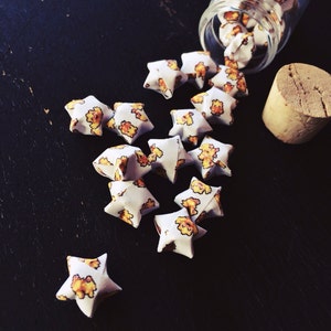May include: A glass jar filled with white origami stars with a yellow and orange pattern. The stars are spilling out of the jar onto a black surface.