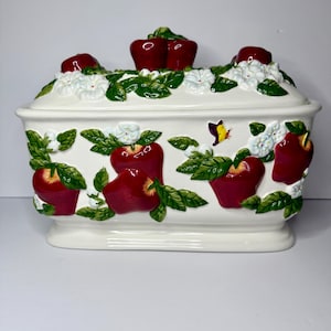May include: A rectangular ceramic lidded container in cream white, featuring a raised relief design of red apples, green leaves, and white blossoms. A small yellow and black butterfly is depicted on the front. The lid has two sculpted red apple handles.