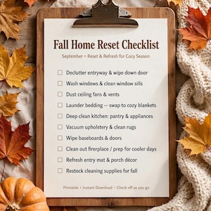 May include: A wooden clipboard holding a white paper titled Fall Home Reset Checklist with a list of tasks including decluttering, cleaning, and refreshing for the season, surrounded by orange autumn leaves and a cream knit fabric.