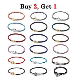 May include: A collection of fifteen braided cord bracelets in various colors including red, blue, black, brown, purple, and pink. Each bracelet features a metallic clasp, some spherical and some cylindrical, in gold or silver tones. Text reads Buy 2, Get 1.