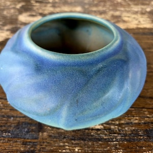 May include: A small, round ceramic bowl with a wide opening and a matte finish. The exterior features a textured, swirling leaf-like pattern in shades of blue and teal, with a smooth, rounded rim and a deep, hollow interior.