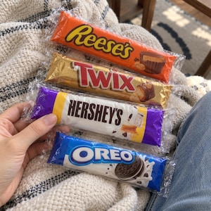 Pode incluir: Quatro barras de chocolate retangulares em embalagens plásticas transparentes empilhadas verticalmente. A barra superior é laranja da Reese's, seguida por uma Twix dourada, uma Hershey's branca e roxa e uma barra de chocolate Oreo azul.