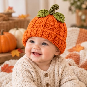 May include: A bright orange crocheted beanie hat featuring a ribbed texture and a green crocheted pumpkin stem with two green leaves attached to the top. The hat is worn by a smiling infant dressed in a cream-colored cable knit sweater.