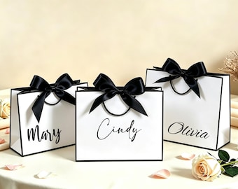 Pode incluir: Três sacos de presente brancos com bordas pretas e laços de fita de cetim preta. Cada saco possui uma alça de cordão preto e um nome personalizado impresso em letra cursiva preta: Mary, Cindy e Olivia. Os sacos estão em fila.