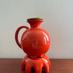 May include: A bright orange ceramic pitcher with a spherical body, a curved handle, and a flared neck, topped with a light-colored circular lid. The pitcher rests on a matching orange ceramic base featuring multiple arched legs.