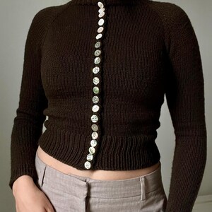 Könnte beinhalten: Ein dunkelbrauner, langärmeliger Strickpullover mit hohem Stehkragen und einer vertikalen Reihe kleiner, runder, schimmernder weißer Knöpfe auf der Vorderseite. Der Pullover hat gerippte Bündchen am Saum und an den Ärmeln.