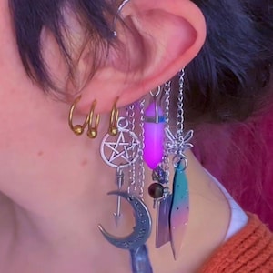 May include: A collection of earrings featuring a variety of charms and pendants. The earrings include gold hoops, a silver pentagram, a crescent moon, and a purple crystal. Additional charms include a dragonfly and a gradient-colored feather.