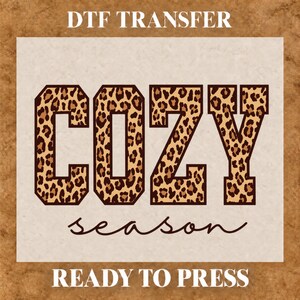 May include: A DTF transfer sheet featuring the word COZY in large block letters with a brown and black leopard print pattern, followed by the word season in a cursive script font, with the text DTF TRANSFER and READY TO PRESS.