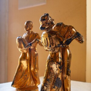 May include: Two gold-painted figurines of a couple dancing. The woman is wearing a long dress and the man is wearing a suit. The figurines are standing on a white surface.