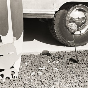 Op de afbeelding: Zwart-wit afbeelding met een decoratief metalen alienfiguur, een surfplank en een vintage voertuigband. De alien staat op een bed van kleine rotsen. De band heeft een chromen wieldop.