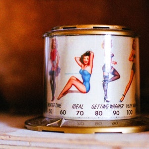 May include: A white metal cylinder with a gold top and bottom. The cylinder has a thermometer scale with numbers from 50 to 100. The scale is labeled "Neater Time", "Ideal", "Getting Warmer", and "Very". The cylinder has a graphic of four women in various poses.