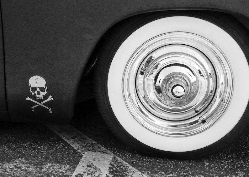 May include: Monochrome image of a vintage car wheel. The tyre features a white sidewall and a shiny chrome hubcap. A skull and crossbones emblem is displayed on the vehicle's body.