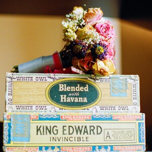 Könnte beinhalten: Ein Strauß aus rosa, weißen und gelben Blumen mit grünen Blättern liegt auf zwei Schachteln. Die obere Schachtel ist weiß mit dem Text "White Owl Blended with Havana" darauf gedruckt. Die untere Schachtel ist blau und weiß mit dem Text "King Edward Invincible" darauf gedruckt.