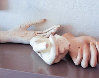 May include: Three white plaster mannequin hands arranged on a brown surface. The hands are in various poses, with one hand reaching out, one hand cupped, and one hand closed into a fist.