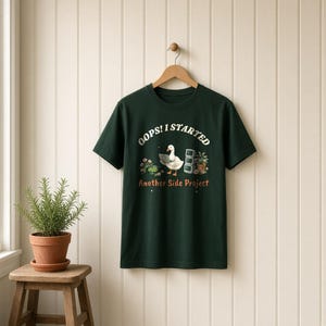 May include: A dark green short-sleeve t-shirt featuring a graphic of a white duck using a laptop. The text reads OOPS! I STARTED Another Side Project. The design includes a small potted plant, a stack of monitors, and a bowl of green peas.