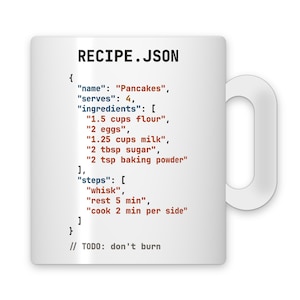 May include: A white ceramic mug featuring a black, blue, and red JSON code snippet titled RECIPE.JSON. The code lists ingredients for pancakes, including flour, eggs, milk, sugar, and baking powder, with steps to whisk, rest, and cook, plus a comment.
