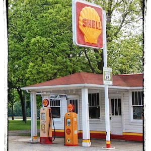 May include: A vintage Shell gas station with a red and yellow sign featuring the Shell logo. The station has two gas pumps, a white building with a red roof, and a sign that reads "Historic Route 66".