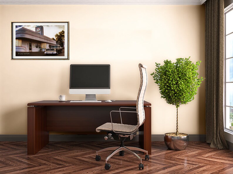 May include: A home office with a brown wooden desk, a white office chair, a computer monitor, a potted plant, and a framed picture of a vintage diner with a car parked in front.