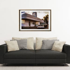 May include: A vintage black car parked in front of a retro diner with a red and white sign that says "Diner". The diner has a large window and a white awning. The image is in a gold frame.