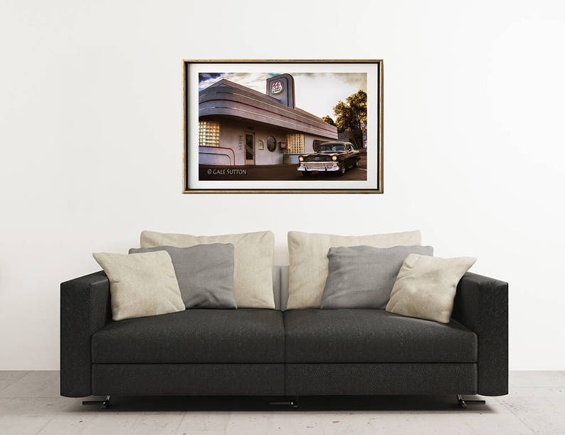 May include: A vintage black car parked in front of a retro diner with a red and white sign that says "Diner". The diner has a large window and a white awning. The image is in a gold frame.