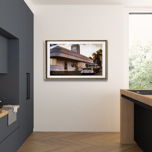 May include: A framed photograph of a vintage diner with a classic car parked in front. The diner has a white exterior with a red and blue sign that says "Diner". The car is a dark green 1950s model with chrome accents. The photograph is set against a white wall in a modern kitchen.