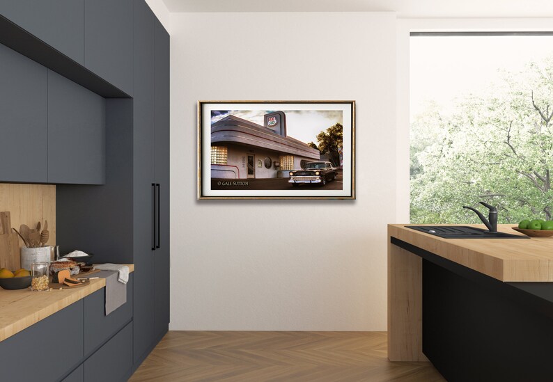 May include: A framed photograph of a vintage diner with a classic car parked in front. The diner has a white exterior with a red and blue sign that says "Diner". The car is a dark green 1950s model with chrome accents. The photograph is set against a white wall in a modern kitchen.