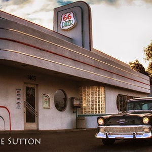 May include: A black classic car parked in front of a retro diner with a "66 Diner" sign. The diner has a white and gray exterior with red accents and a large glass block window.