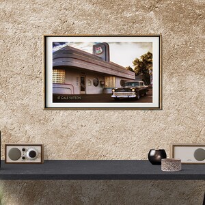 May include: A vintage black car parked in front of a retro diner with a red and white sign that says "Diner". The diner has a white exterior with a gray trim and large windows. The photo is framed in a gold frame.