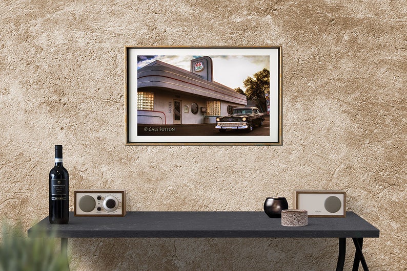 May include: A vintage black car parked in front of a retro diner with a red and white sign that says "Diner". The diner has a white exterior with a gray trim and large windows. The photo is framed in a gold frame.