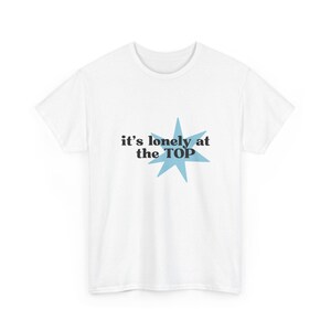Puede incluir: Una camiseta blanca de manga corta con un gráfico de estrella azul claro detrás del texto negro que dice, it's lonely at the TOP. La prenda cuenta con un cuello redondo y un corte estándar para un aspecto sencillo y limpio.