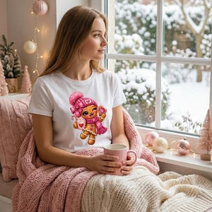 Op de afbeelding: Een wit t-shirt met korte mouwen en een afbeelding van een glimlachende peperkoekfiguur met een roze gebreide muts en sjaal, die een roze mok en een hartvormig koekje vasthoudt, met roze wangen en een roze strik.