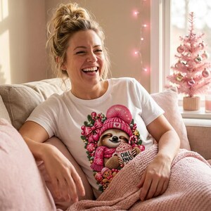 K&ouml;nnte beinhalten: Ein wei&szlig;es T-Shirt mit einer Grafik eines Faultiers, das eine rosa Strickm&uuml;tze und einen Schal tr&auml;gt und eine Tasse mit der Aufschrift CHILL XMAS h&auml;lt. Das Design enth&auml;lt rosa Ornamente, eine Schleife und einen Lebkuchenmann.