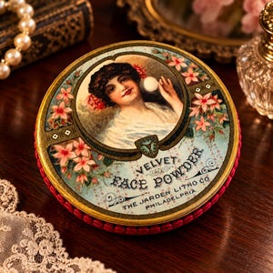 May include: A round metal tin for Velvet Face Powder by The Jarden Litho Co Philadelphia. The lid features a central illustration of a person with dark hair and red flowers, surrounded by pink floral motifs on a light blue background.