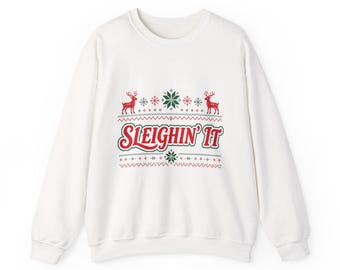 May include: A white long-sleeve sweatshirt featuring a festive graphic print in red and green. The design includes two reindeer, snowflakes, and the text "SLEIGHIN' IT" centered on the chest, surrounded by decorative geometric patterns.