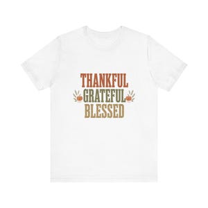Puede incluir: Una camiseta blanca de manga corta con el texto THANKFUL GRATEFUL BLESSED en líneas apiladas. La palabra THANKFUL es naranja, GRATEFUL es verde y BLESSED es color canela, flanqueada por pequeñas calabazas naranjas y espigas.