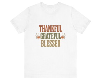 Peut inclure: Un t-shirt blanc à manches courtes avec le texte THANKFUL GRATEFUL BLESSED écrit en lignes superposées. Le mot THANKFUL est orange, GRATEFUL est vert et BLESSED est beige, entouré de petites citrouilles orange et d'épis de blé.