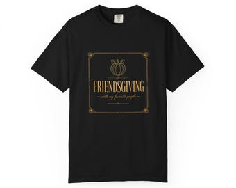 May include: A black short-sleeve t-shirt featuring a gold-colored graphic print. The design includes a pumpkin icon, a decorative border, and the text "FRIENDSGIVING with my favorite people" centered on the chest of the garment.