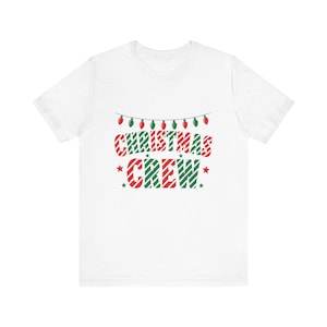 May include: A white short-sleeved t-shirt featuring a graphic print of a string of red and green Christmas lights above the text CHRISTMAS CREW in red and green diagonal stripes, accented by small green stars on the sides of the word CREW.
