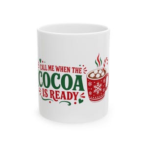 May include: A white ceramic mug featuring the red and green text "CALL ME WHEN THE COCOA IS READY" alongside an illustration of a red mug filled with cocoa, marshmallows, and a candy cane, decorated with white snowflakes and green hearts.